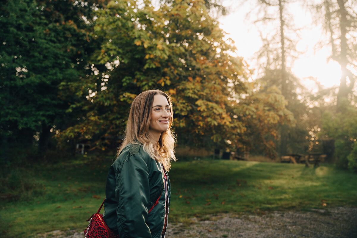 En kvinna ler och tittar upp mot himlen medan hon står i en park eller skog.